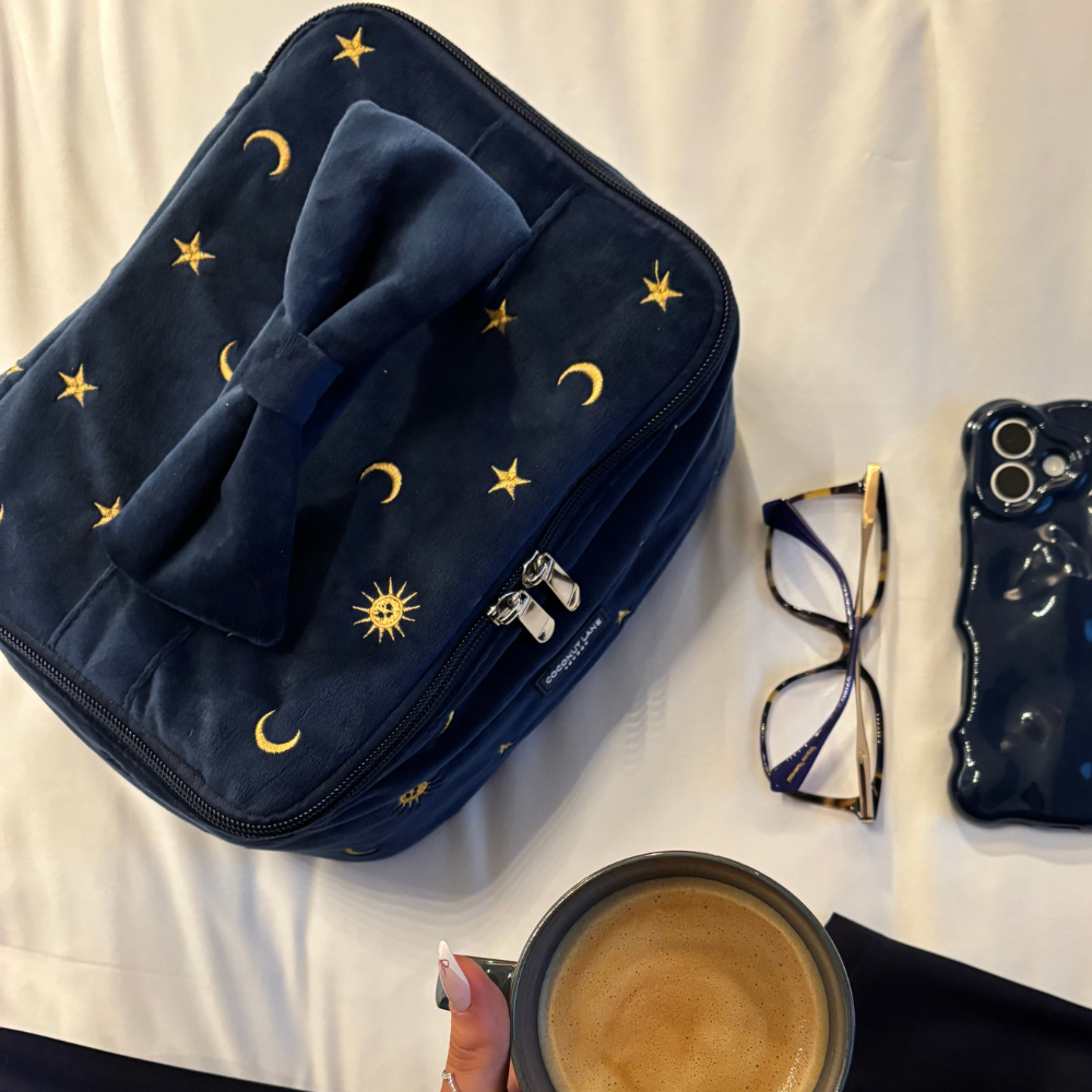 Navy celestial travel vanity case by Coconut Lane on a white surface, with glasses, a phone, and a coffee nearby.