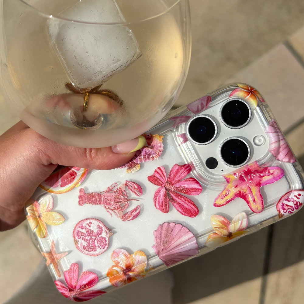 Hand holding a wine glass and phone with Coconut Lane clear case, Rosé Coast design with seashell and flower pattern.