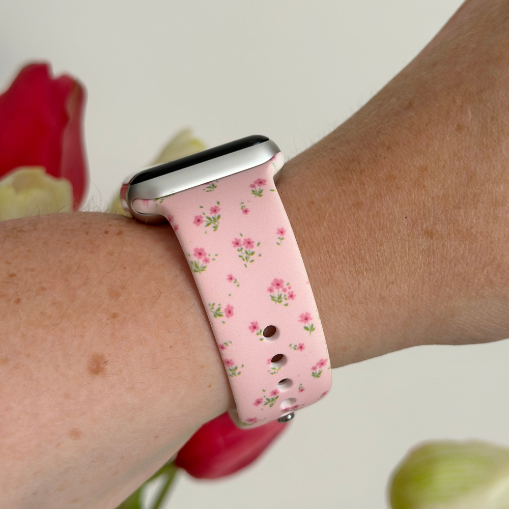 Pink floral Apple Watch strap on wrist, Coconut Lane branding, blurred red and white flowers in the background.