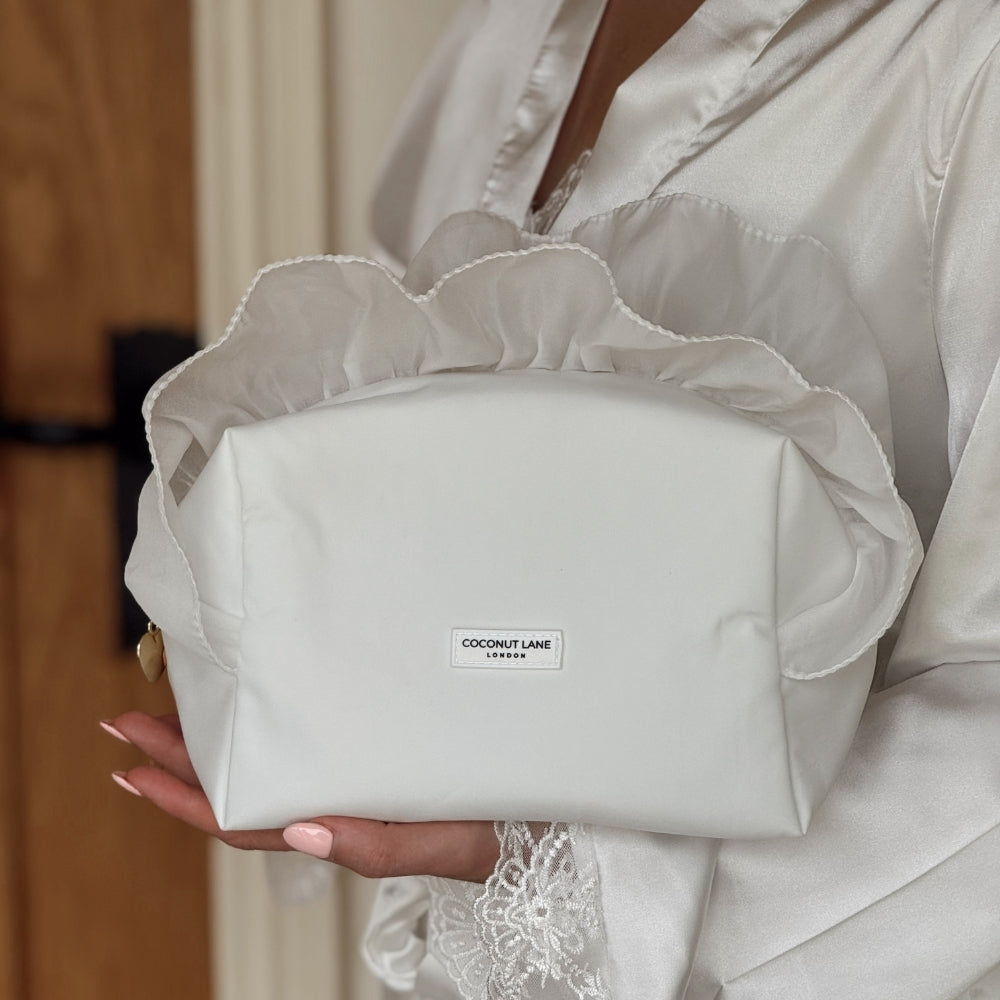 bride in satin dressing gown holding a White Ruffle Satin Makeup Bag with 'Coconut Lane' branding.
