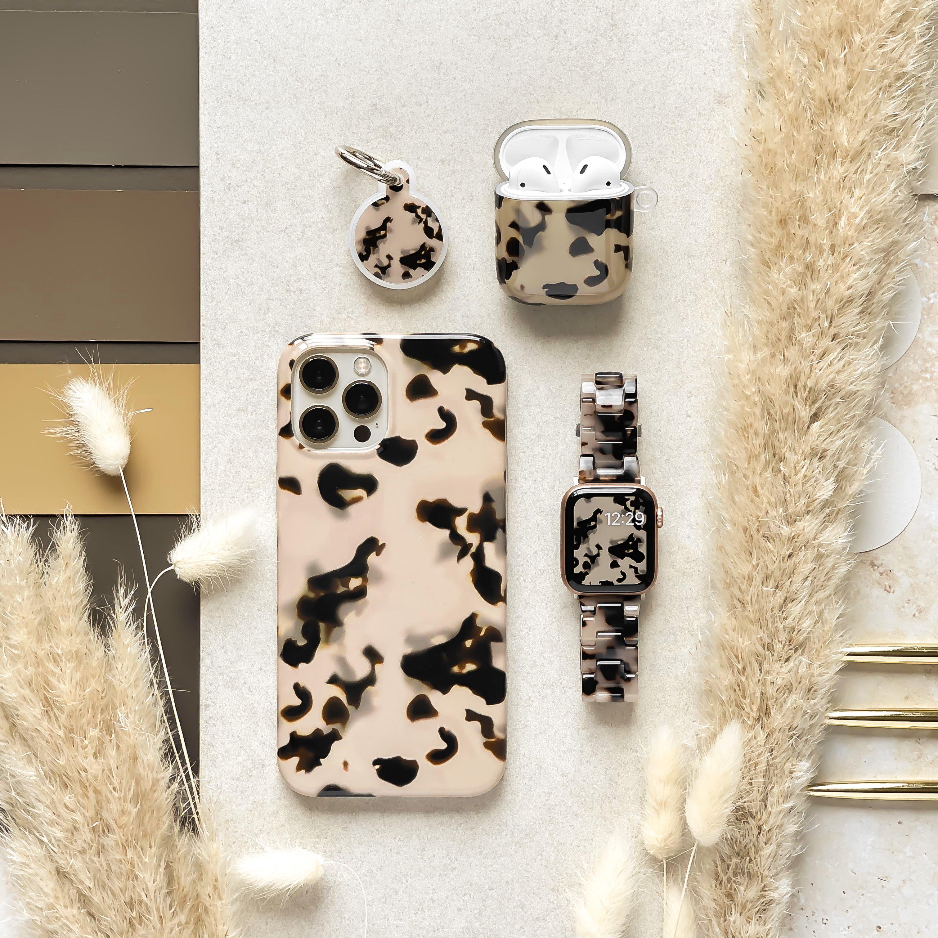 A beige background featuring a Luxe Ivory Tort Apple Watch Strap from Coconut Lane, along with a matching phone case, AirPods case, watchband, and keychain in beige and black abstract patterns. Beige and black sample cards and dried pampas grass are arranged around, enhancing the aesthetic presentation.