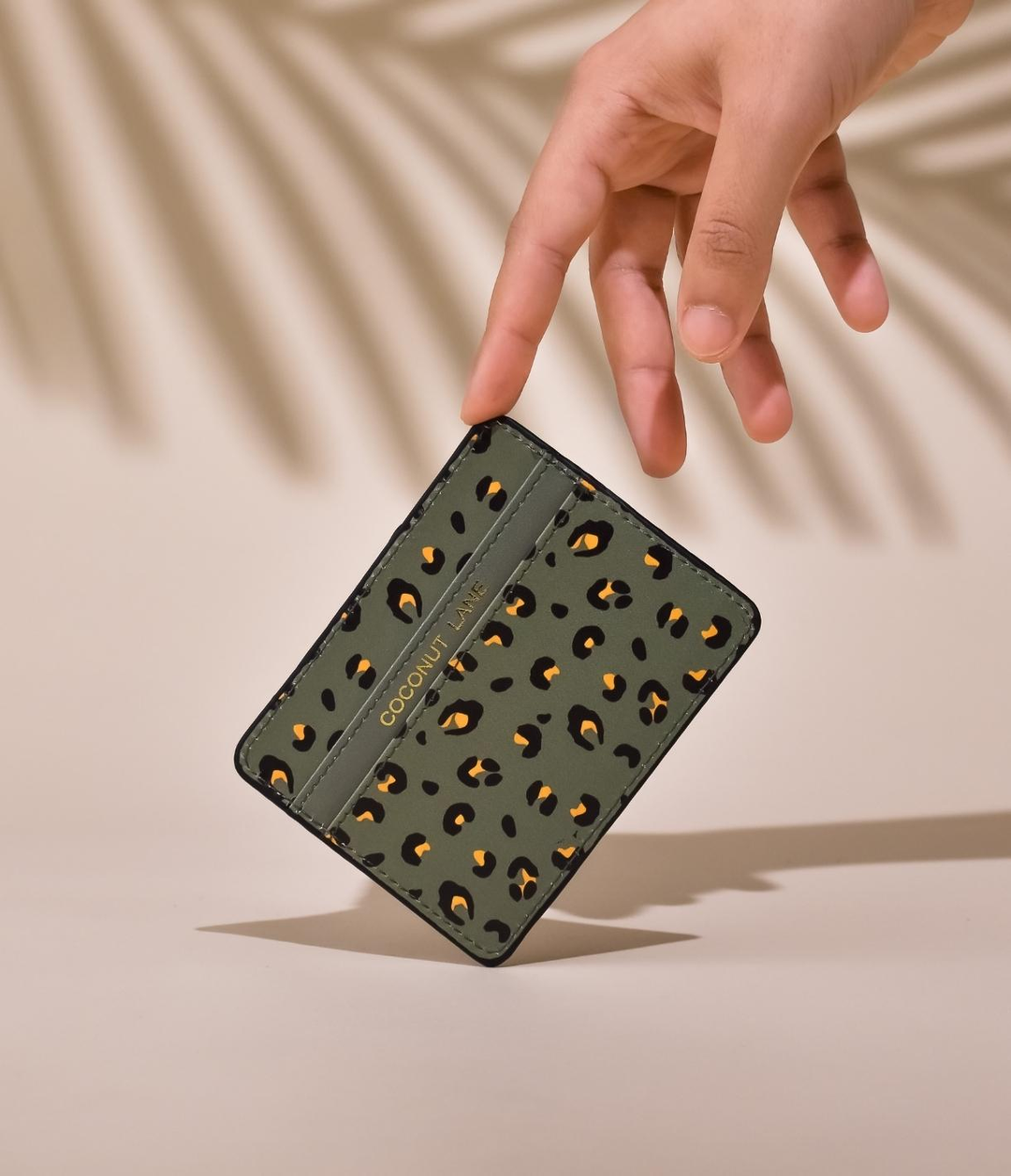 A hand holds a stylish cardholder with a leopard print design against a beige background with palm shadows.