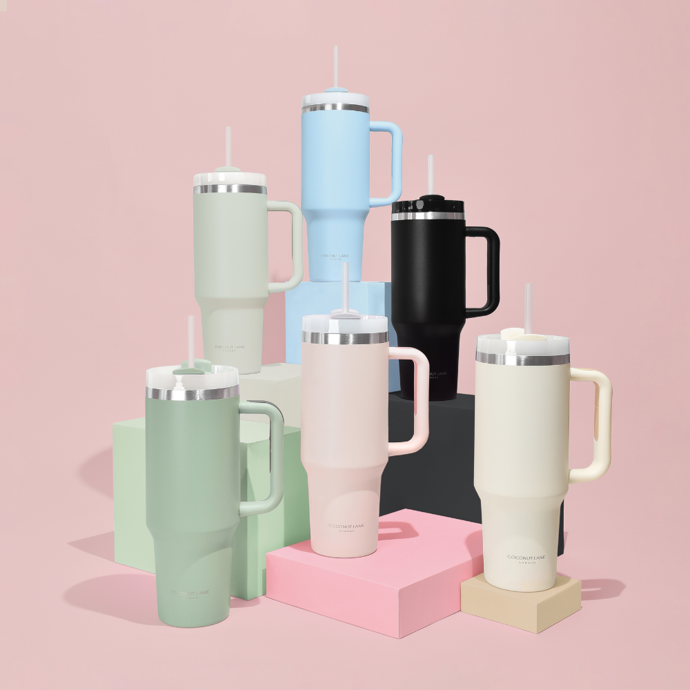 Six colorful, reusable water tumblers with handles and straws are arranged on pastel blocks against a light pink background. The tumblers come in soft shades of green, beige, pink, blue, black, and cream.