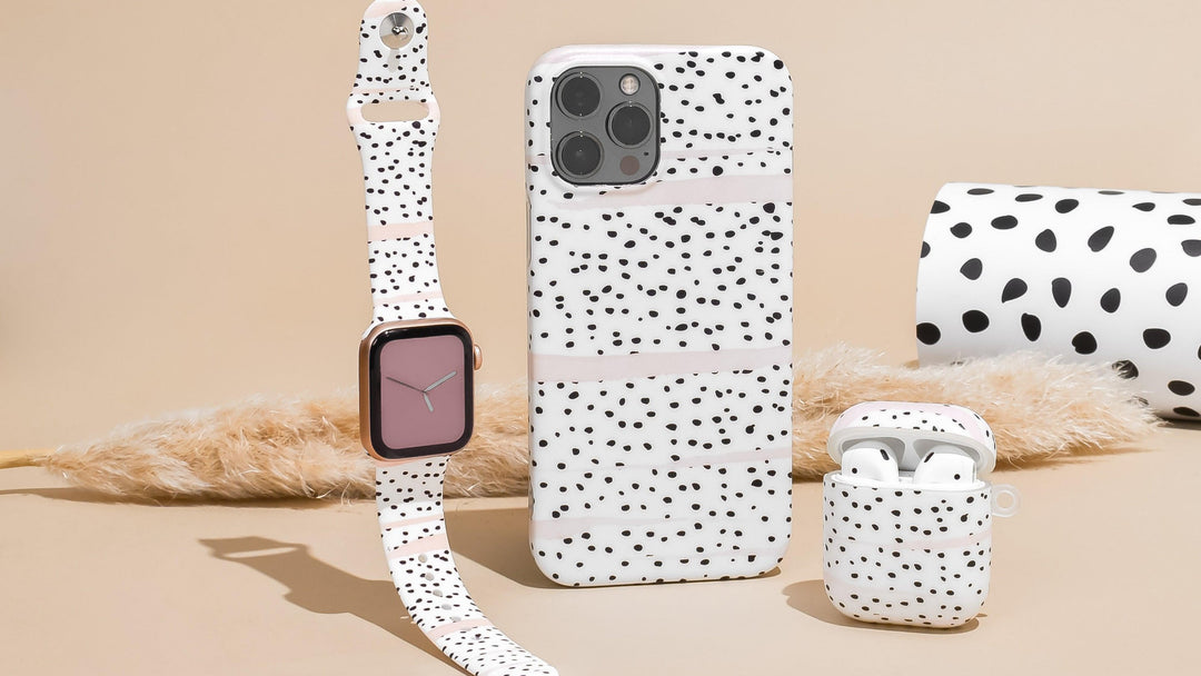 A smartphone case, smartwatch strap, and wireless earbud case with a white and black speckled pattern are arranged on a beige surface. A roll of the same patterned wrapping paper and a dried grass stem are nearby.