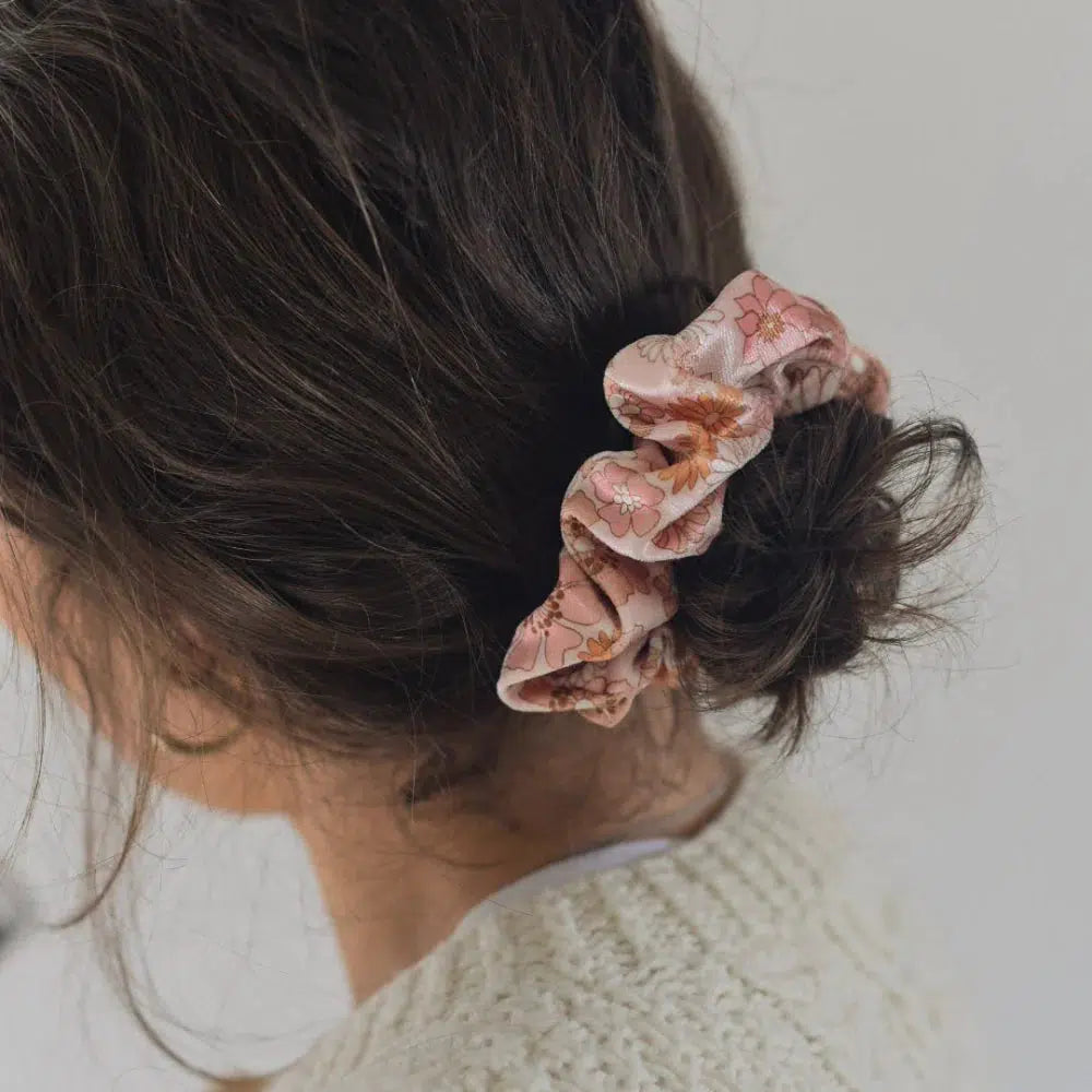A woman with dark hair styled in a messy bun, secured by a pink floral scrunchie. She is wearing a cream-colored sweater and is turned slightly away from the camera, set against a plain background.