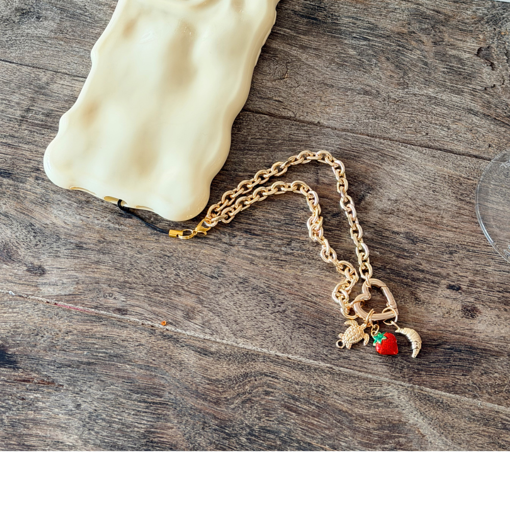A gold chain necklace with a heart pendant, crescent moon, flower, and star charms. "THE CHARM STUDIO" text appears in black in the lower right corner on a white background.
