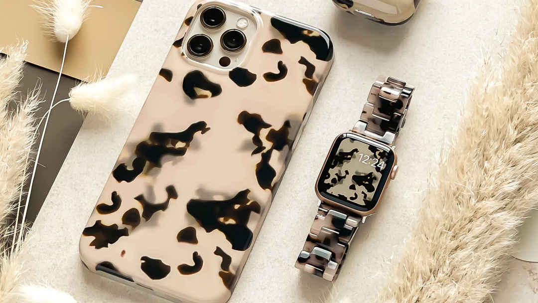 A smartphone and smartwatch with matching abstract camo-patterned covers are placed on a light surface, surrounded by dried grass decor. The smartwatch displays the time as 12:24.