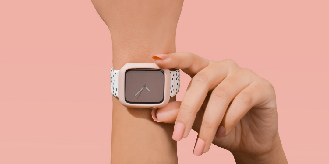 Close-up of a smartwatch with a blank screen on a wrist against a soft pink background. A hand with pink nail polish is touching the side of the watch. The watch has a white band with a subtle pattern.