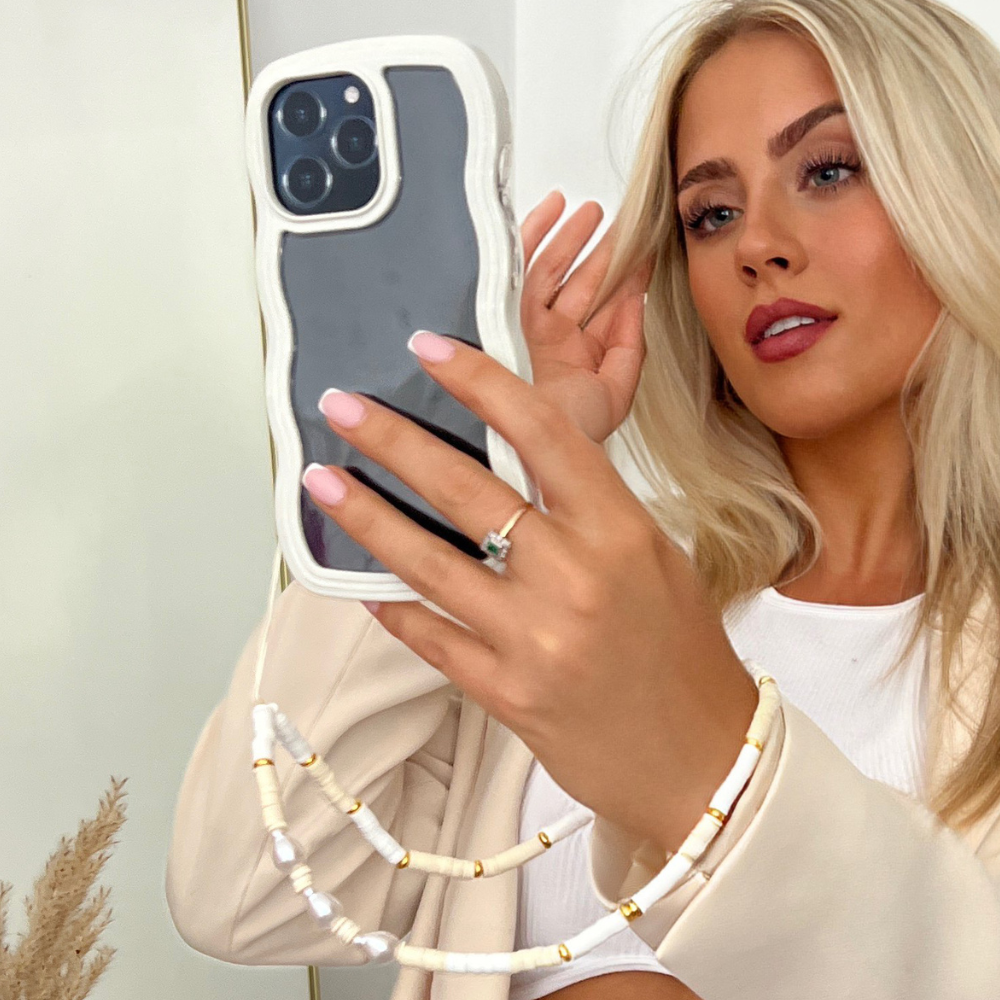 A woman with long blonde hair takes a selfie with a white iPhone enclosed in a wavy white case. She’s dressed in a neutral-toned outfit, showcasing a ring on her finger and wearing the Beaded Phone Strap - Nude & Pearl by Coconut Lane on her wrist. A stylish phone charm dangles from the device, adding flair, as dried pampas grass appears in the background.
