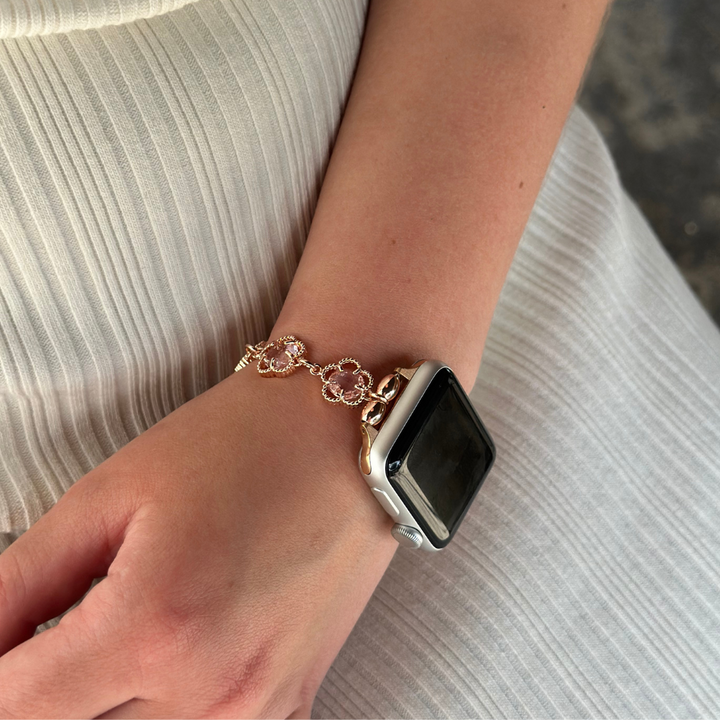 A close-up of a person's wrist adorned with the Coconut Lane Rose Gold Flower Bracelet Apple Watch Strap, featuring intricate links and floral designs. The person is wearing a white, pleated dress. The background is blurred, highlighting the wrist and bracelet.