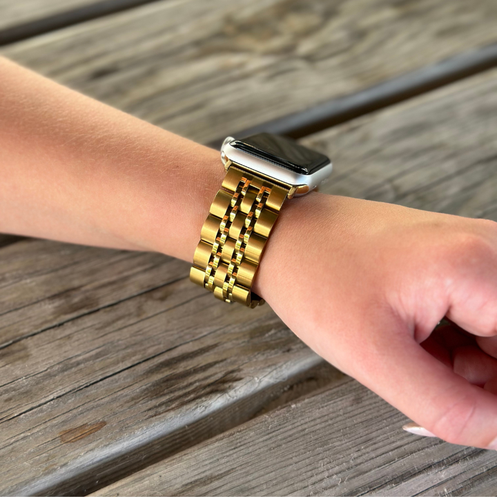 Close-up of a person's wrist wearing the Coconut Lane Stainless Steel Apple Watch Strap in Gold, featuring a rectangular digital screen. The wrist is resting on a weathered wooden surface, with the hand relaxed and fingers slightly curled.