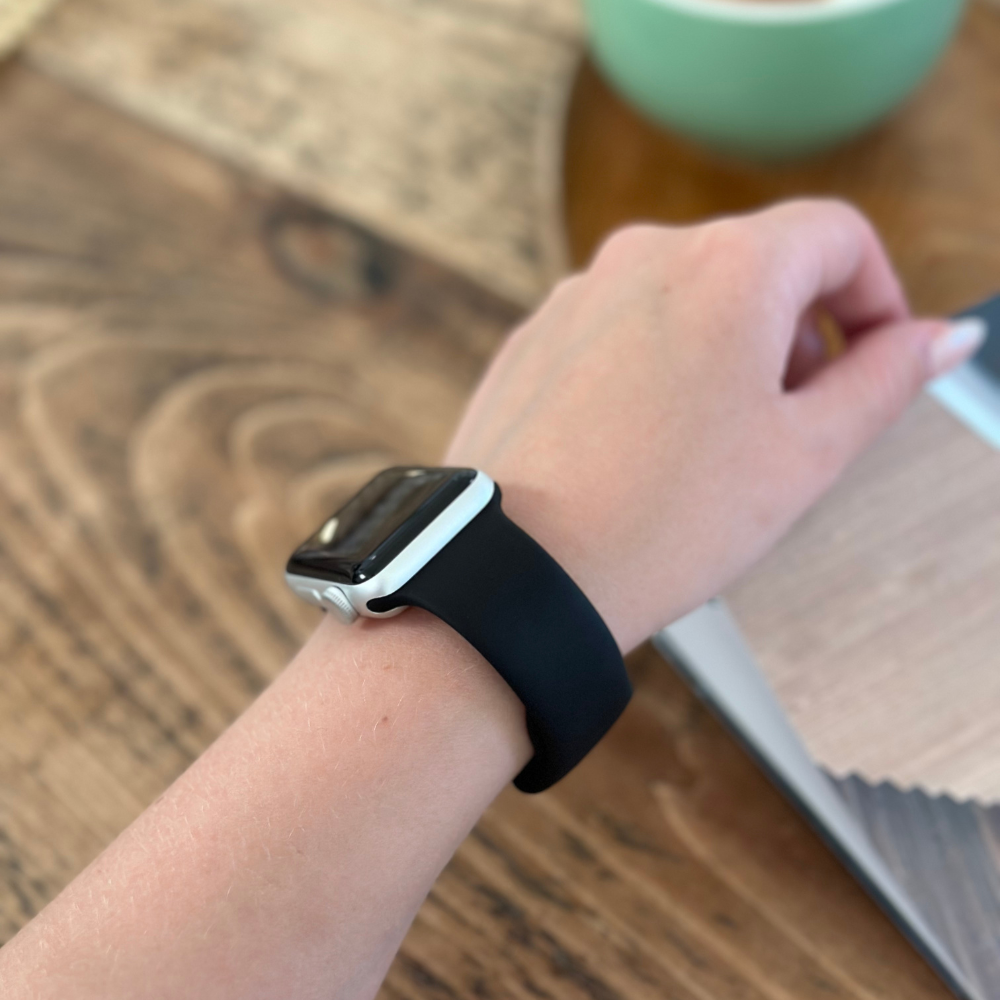 Close-up of a person's left wrist wearing a Coconut Lane NAKD Apple Watch Strap - Midnight Black. The person is seated at a wooden table, with an open book or magazine and a green cup partially visible in the background.
