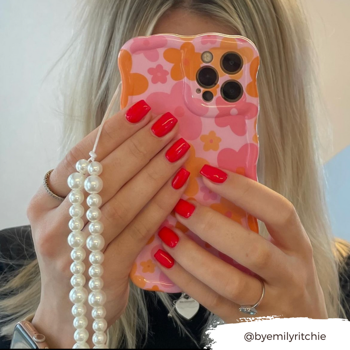 A person with blonde hair is holding a smartphone in front of their face, which is protected by Coconut Lane's Wavy Phone Case - Pink & Orange Bloom. The case features a floral design with pink and orange flowers and a string of pearls. The person has bright red nail polish. The Instagram handle "@byemilyritchie" is shown in the bottom right corner.