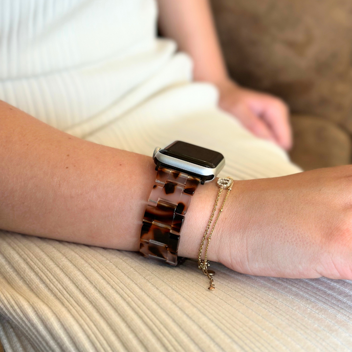 A person is shown wearing an Apple Watch with a Luxe Ivory Tort Apple Watch Strap from Coconut Lane and a delicate gold bracelet on their wrist. They are seated, dressed in a light-colored, textured fabric dress, with their arm resting on their lap. The background features a blurred brown surface.