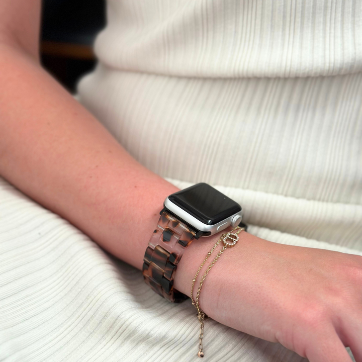 Close-up of a person's wrist displaying a Coconut Lane Luxe Ivory Tort Apple Watch Strap alongside a delicate, gold-chain bracelet. The individual is dressed in a white, ribbed fabric outfit with their arm resting on their lap.