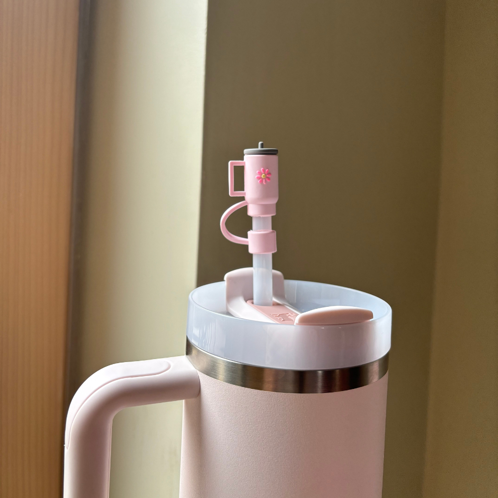 A close-up of Coconut Lane's Mini Pink Tumbler Straw Cover, featuring a silicone spout button for controlling the flow of liquid. The tumbler is set against a beige background, with ambient light adding a warm tone to the scene, and includes a charming matching straw topper.