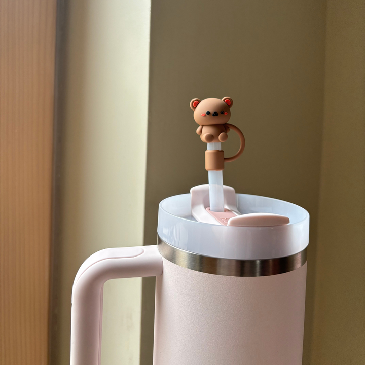 A white tumbler with a handle and lid features the Teddy Bear Straw Cover from Coconut Lane, adorned with a small, cute silicone bear-shaped decoration. The tumbler is positioned against a neutral-colored wall, gently illuminated by soft natural light.