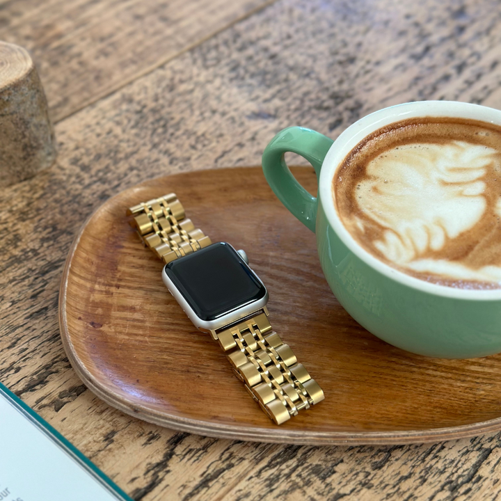 A Stainless Steel Apple Watch Strap - Gold by Coconut Lane lies on a wooden tray beside a green cup filled with latte art coffee on a wooden table. The tabletop also includes part of another item at the top-left corner, possibly a small container or jar.