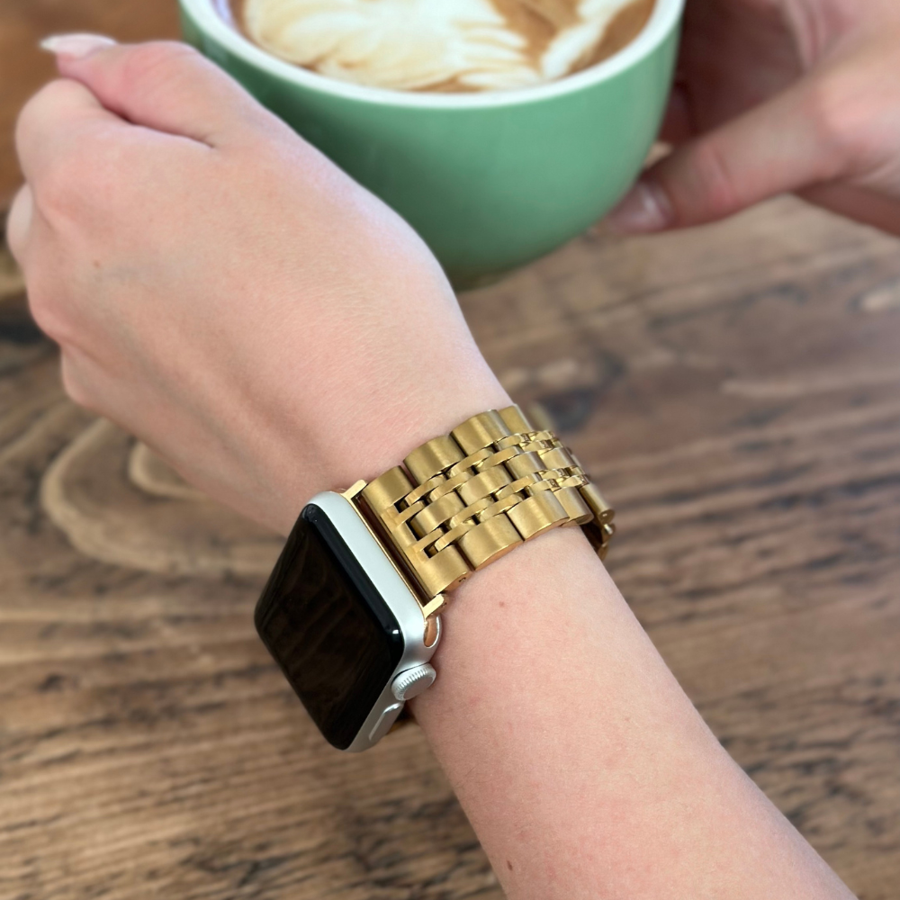 A person is holding a light green cup of cappuccino with latte art, their wrist adorned with Coconut Lane's Stainless Steel Apple Watch Strap in gold, on a wooden table background.
