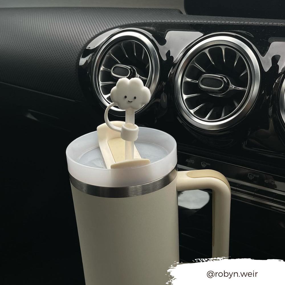 A cream-colored travel mug with a handle sits in a car's cup holder. The Coconut Lane mug boasts a lid adorned with the White Cloud Straw Cover, featuring a charming smiling cloud figurine on top. The background reveals the car’s dashboard with air vents, while the text "@robyn.weir" appears at the bottom right.