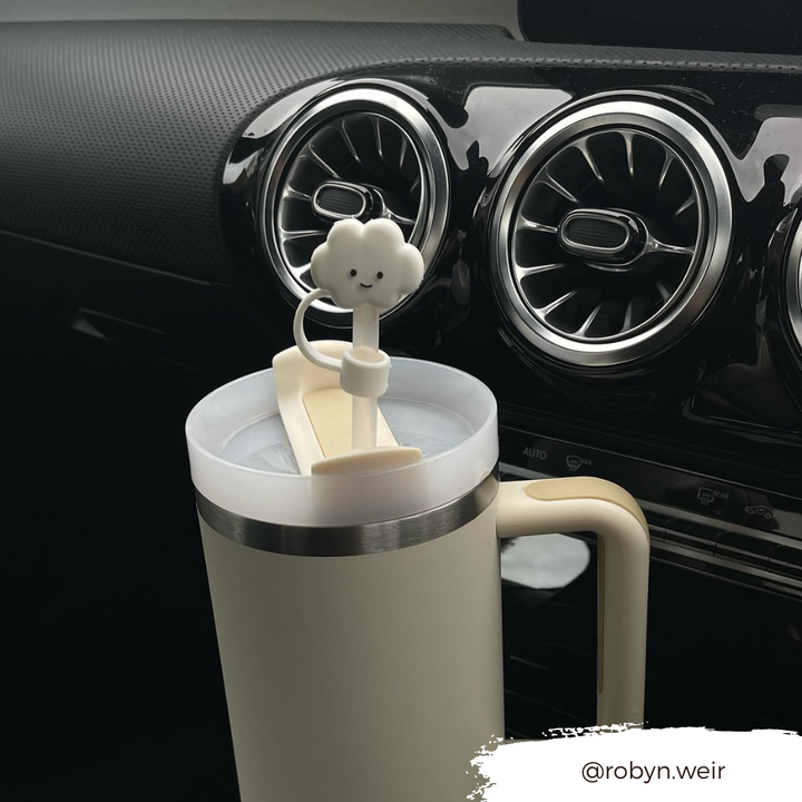 A cream-colored travel mug with a handle sits in a car's cup holder. The Coconut Lane mug boasts a lid adorned with the White Cloud Straw Cover, featuring a charming smiling cloud figurine on top. The background reveals the car’s dashboard with air vents, while the text "@robyn.weir" appears at the bottom right.