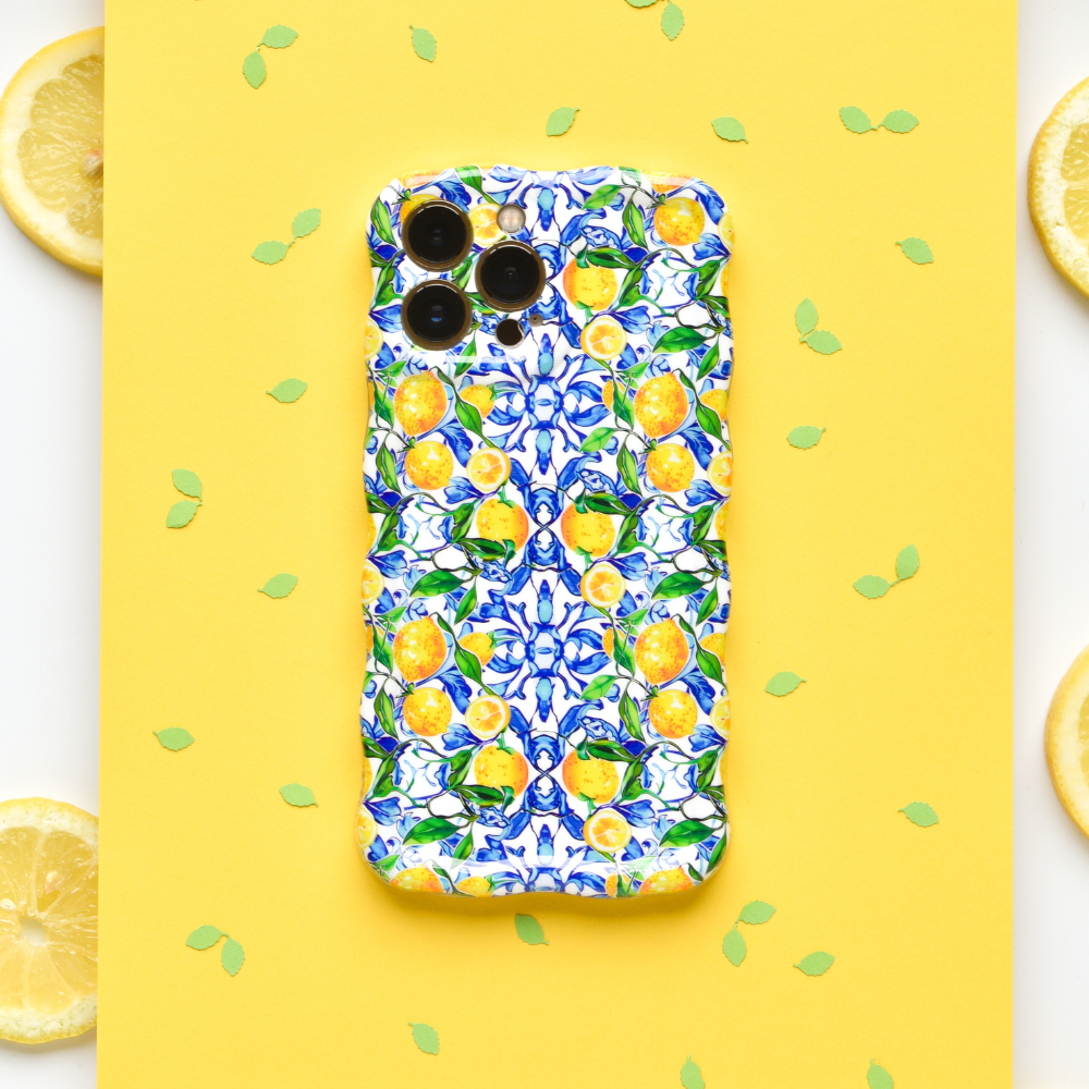 The Wavy Phone Case - Amalfi Lemon by Coconut Lane is showcased on a yellow background. Small green leaf confetti and three lemon slices are placed around it. The case boasts bright blue flowers and yellow Amalfi lemons in a vibrant pattern, providing extra protection for the smartphone.