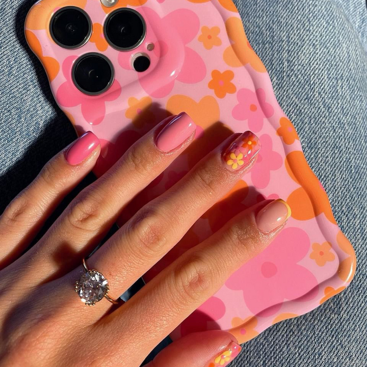 A hand with neatly manicured nails, painted in shades of pink and peach with daisy designs on two nails, holds a smartphone encased in the "Wavy Phone Case - Pink & Orange Bloom" by Coconut Lane. The hand also wears a sparkling diamond ring. The background is a pair of light blue jeans.