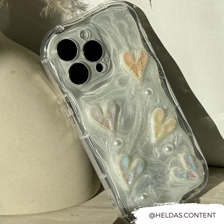 A close-up shot of an iPhone15 enclosed in Coconut Lane's Pearly Hearts Phone Case - Clear, which features sparkling pastel, heart-shaped decorations. The sturdy Pearly Hearts Phone Case lies on a light-colored, textured background. The Instagram handle @HELDAS.CONTENT is displayed in the bottom right corner.