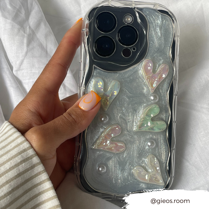 A hand showcasing stylish, swirled orange and white nail art holds an iPhone 15 with the Pearly Hearts Phone Case - Clear from Coconut Lane. This extra protective case features iridescent pearly hearts and a few pearls on a glossy silver background. A white tag at the bottom right corner reads "@gieos.room".