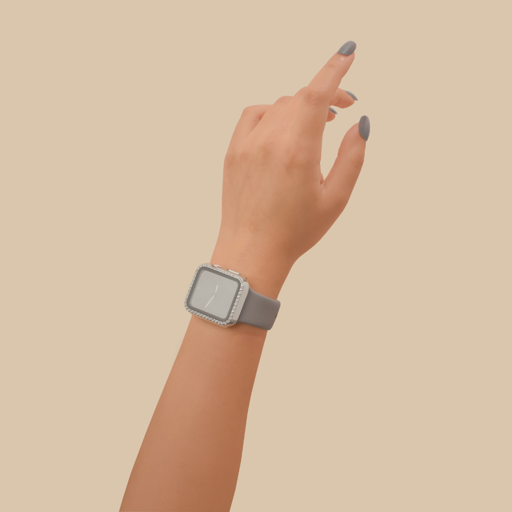 A hand with painted nails wearing a modern Apple Watch with a square face and a gray wristband is positioned against a plain beige background. The fingers are slightly bent as if pointing or touching something, showcasing the elegant 2-in-1 Apple Watch Case & Built-In Screen Protector - Silver Diamante from Coconut Lane that adds a touch of sparkle.