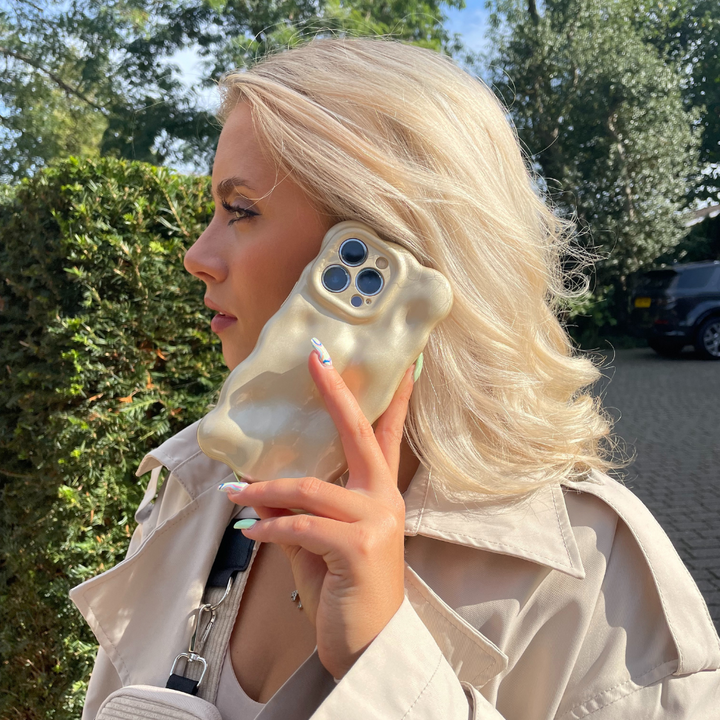 A person with platinum blonde hair holds an iPhone15 with a 3D Pearl Phone Case - Gold by Coconut Lane to their ear. They are wearing a beige trench coat and carrying a small black purse. The background features green foliage and a pathway in a suburban area.