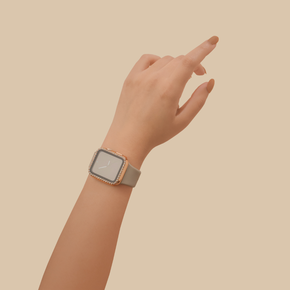 A hand with neutral-toned nails showcases a stylish Coconut Lane 2-in-1 Apple Watch Case & Built-In Screen Protector - Rose Gold Diamante, featuring a beige strap and a sparkly rose gold bezel. The hand is elegantly positioned against a plain, peach-colored background.