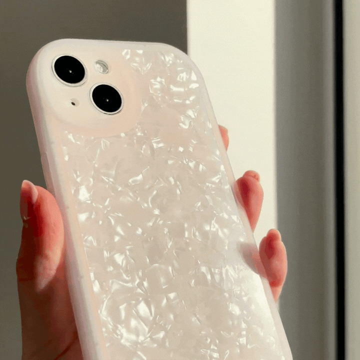 A hand holds a smartphone with a Moonstone Pink Phone Case by Coconut Lane. The phone has two rear camera lenses and is positioned in front of a bright window, emphasizing the glossy finish of the protective phone case.