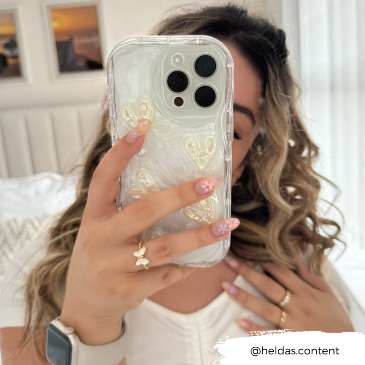 A woman with wavy blonde hair holds up her iPhone15 encased in the Coconut Lane Pearly Hearts Phone Case - Clear, featuring elegant heart designs, taking a selfie. She has floral nail art and wears a ring with a butterfly design. The background includes a white bed and two framed pictures. Text: "@heldas.content".