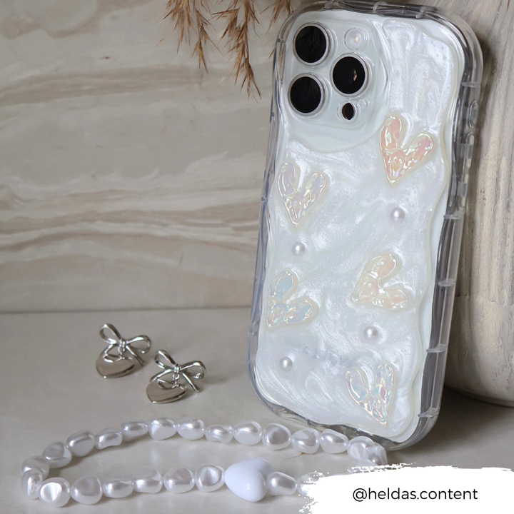 Displayed against a marble backdrop is the Pearly Hearts Phone Case - Clear by Coconut Lane, embellishing an iPhone15 with its pearlescent white casing adorned with shiny heart shapes and pearls. Silver butterfly earrings and a beaded white bracelet accompany the phone. In the bottom right corner, "@heldas.content" is written.