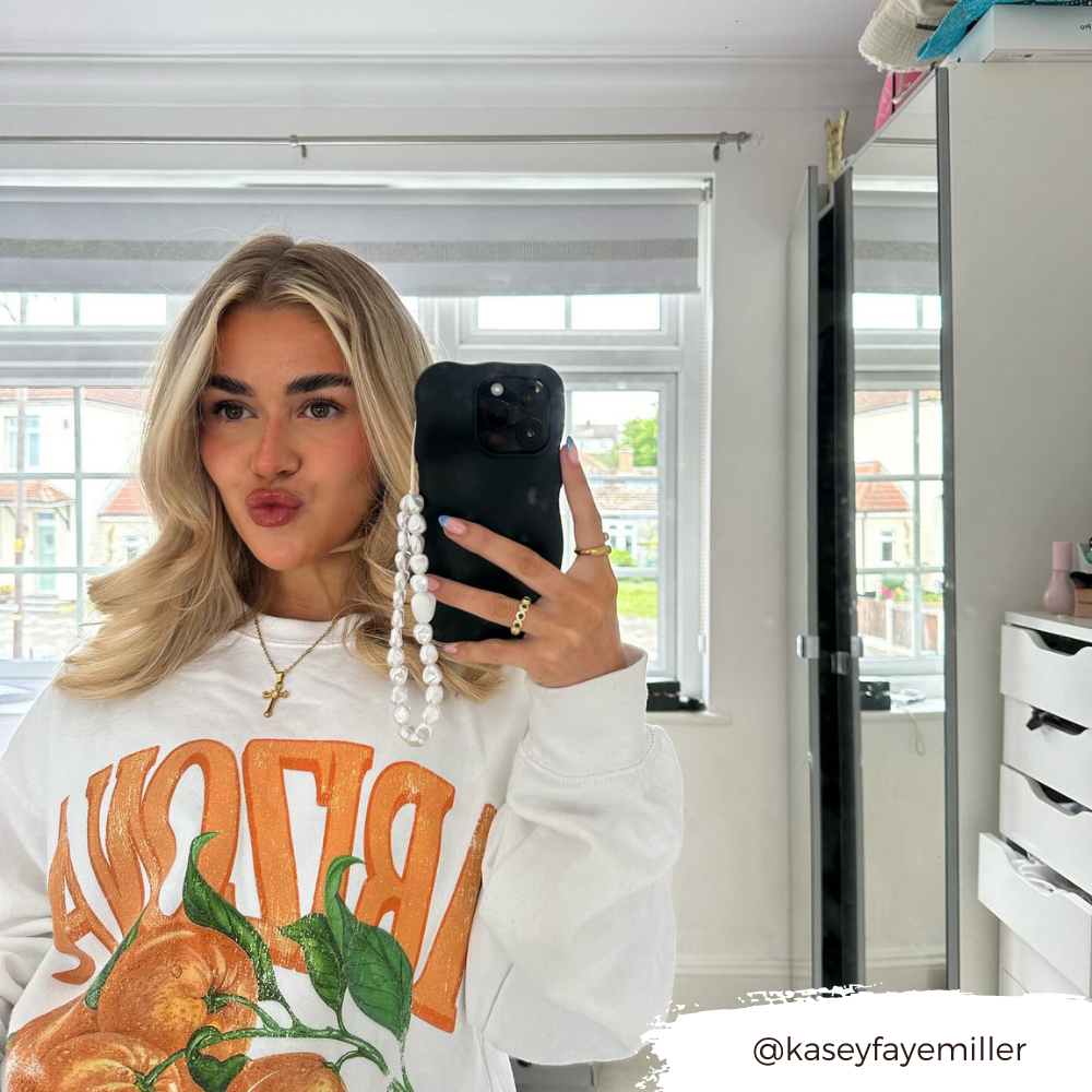 A woman with long blonde hair is taking a mirror selfie in a brightly lit room. She wears a white sweatshirt featuring a bold orange fruit design with the word "BRAVO" on it. Her phone, adorned with the Coconut Lane 3D Boujee Phone Case in an extra protective matte black finish, has a pearl chain attached. A dresser and shelves are visible in the background.