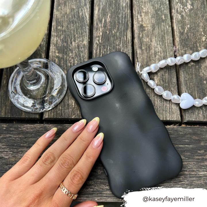 A hand with manicured nails gently touches the Coconut Lane 3D Boujee Phone Case in matte black, displaying clearly visible camera lenses on a wooden surface. Beside it is a beaded bracelet with a white heart charm and a glass of light-colored beverage. Instagram handle @kaseyfayemiller.