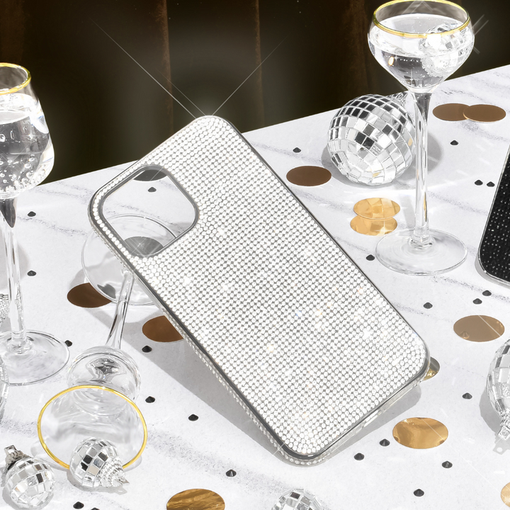 The Disco Sparkle Phone Case - Silver from Coconut Lane, adorned with shimmering rhinestones, rests on a white tablecloth scattered with black and gold confetti. Encircling the case are clear wine glasses and small disco ball ornaments, creating a festive and glamorous atmosphere. Enjoy both style and added protection for your phone.