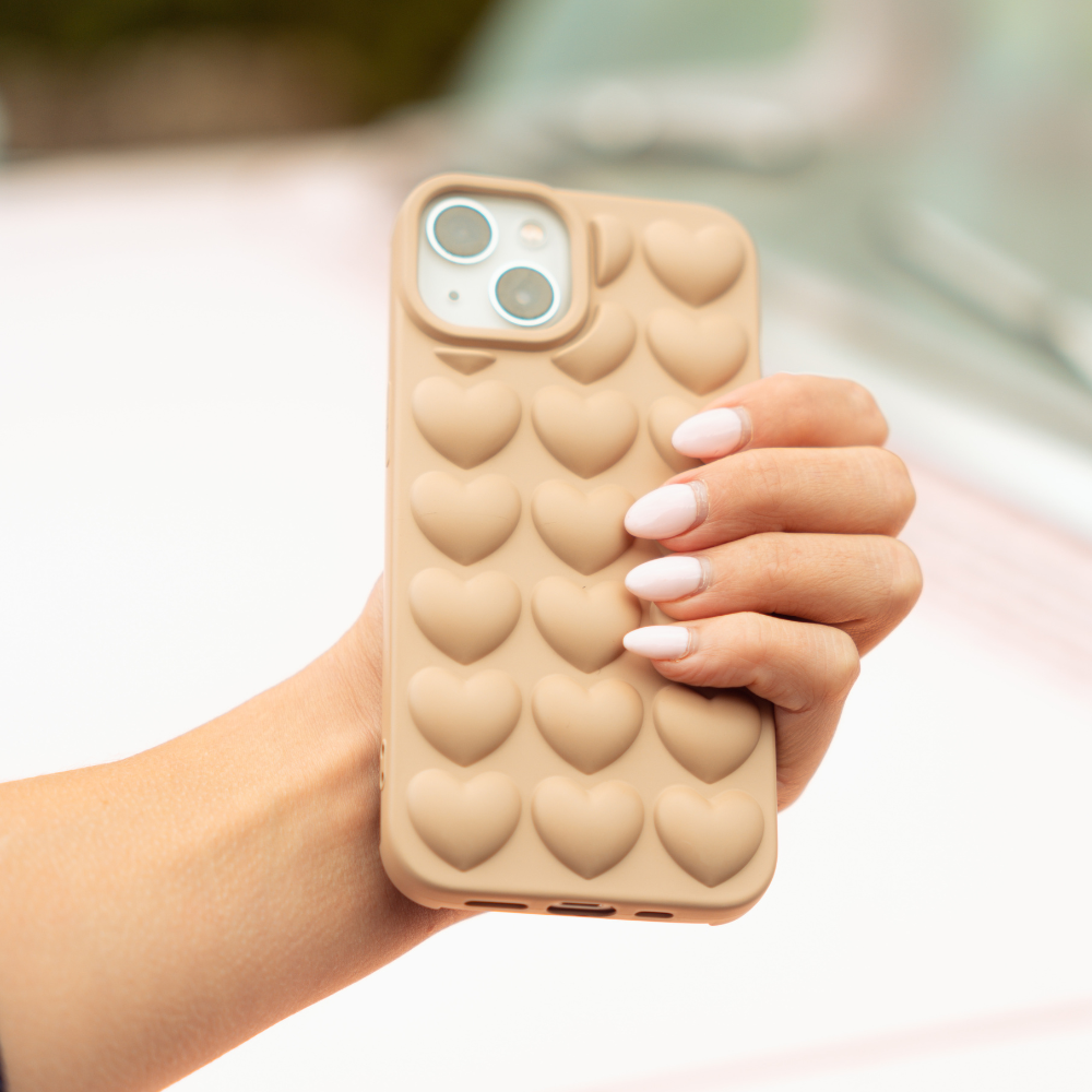 A hand with manicured nails holds an iPhone 15 adorned with the Coconut Lane 3D Hearts Phone Case - Matte Nude. The background is blurred, emphasizing the phone and its chic case.