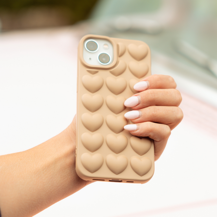 A hand with manicured nails holds an iPhone 15 adorned with the Coconut Lane 3D Hearts Phone Case - Matte Nude. The background is blurred, emphasizing the phone and its chic case.