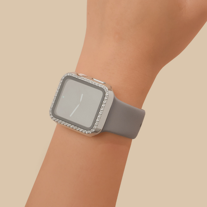 A hand wearing an Apple Watch with a gray strap and a rectangular silver case from Coconut Lane is adorned with small rhinestones around the edge of the screen. The background is a plain light beige color, highlighting the elegant design of this 2-in-1 Apple Watch Case & Built-In Screen Protector - Silver Diamante.