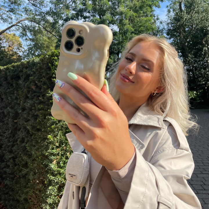 A woman with long blonde hair and mint green nails takes a selfie outdoors. She is wearing a beige trench coat and smiling softly. Her iPhone 15 is adorned with Coconut Lane’s Gold 3D Pearl Phone Case, shaped like a bear, and a small white device is attached to her bag strap. Trees and a hedge are in the background.