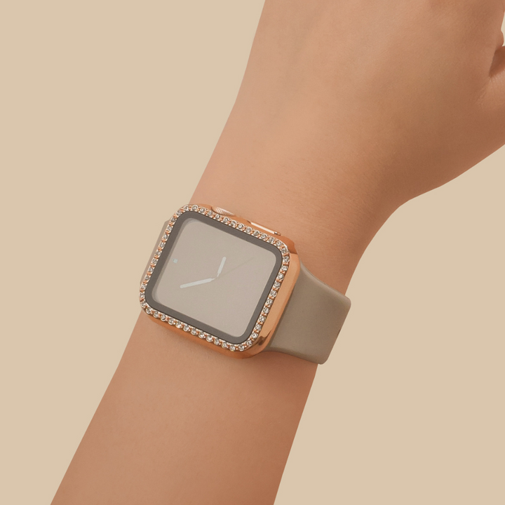A close-up of a wrist wearing the Coconut Lane 2-in-1 Apple Watch Case & Built-In Screen Protector - Rose Gold Diamante, featuring a rectangular face. The watch case has a rose gold diamante frame adorned with small, sparkling stones around the edges and is paired with a light gray strap. The background is a solid beige color.