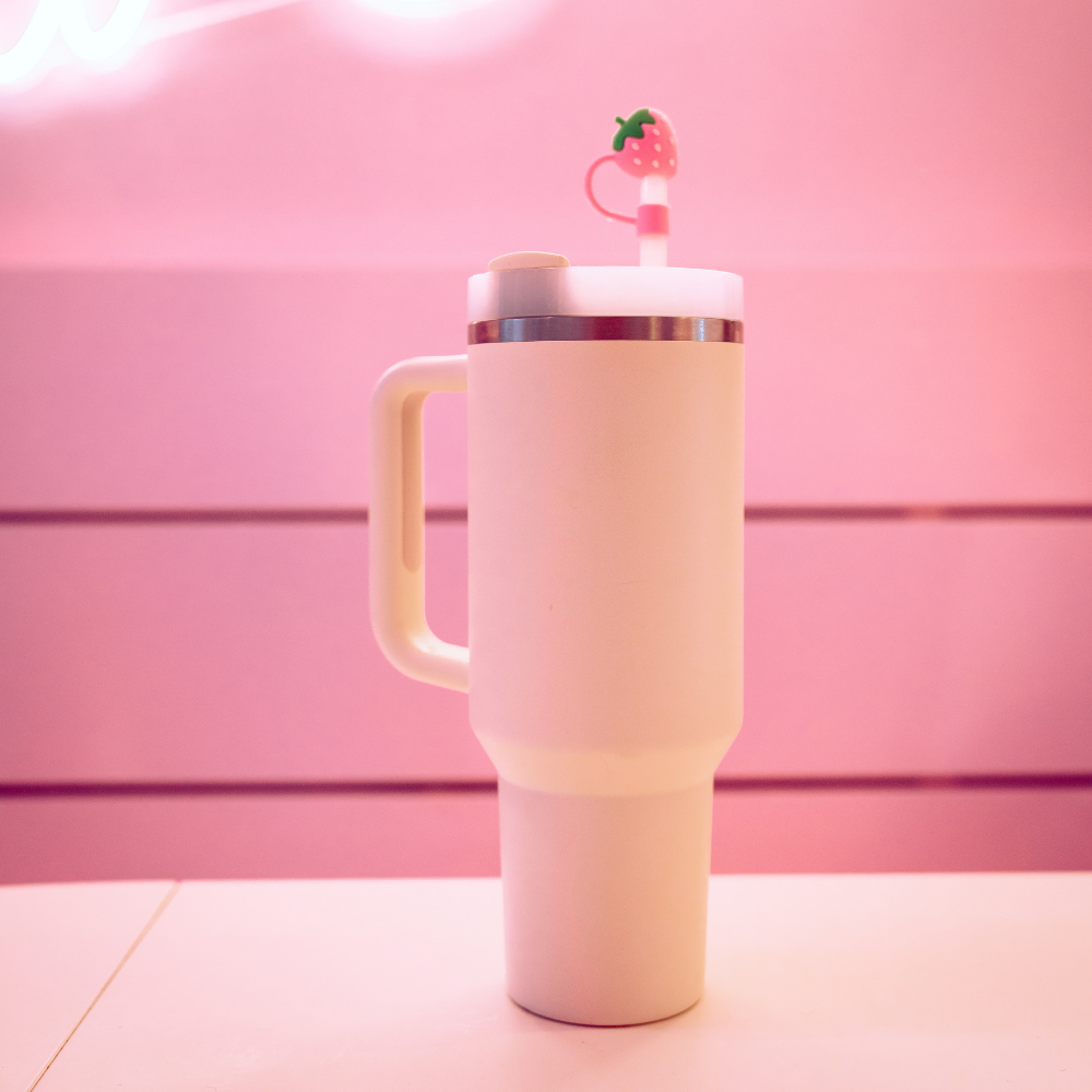 A tall, white travel mug with a handle sits on a pink surface. The mug features a lid adorned with Coconut Lane's Strawberry Straw Cover, and the background is pink, matching the overall aesthetic.