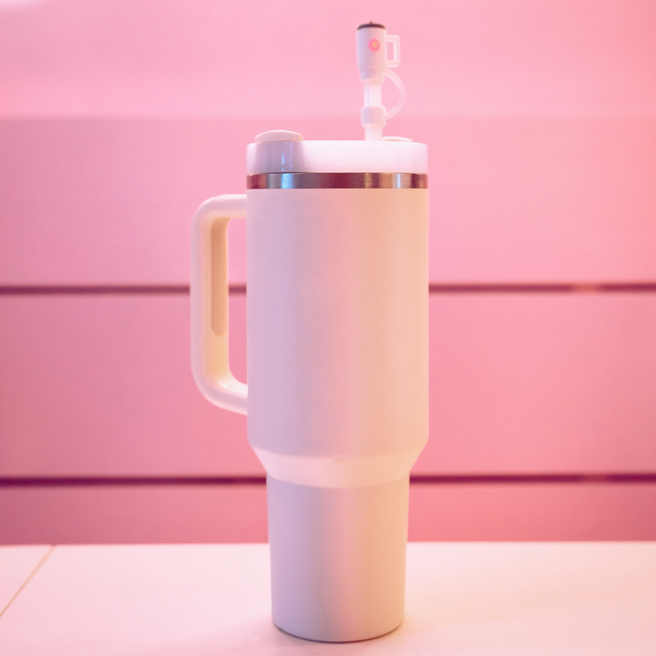 A large Coconut Lane Mini White Tumbler Straw Cover with a handle and a stainless steel rim is centered in the image against a pink background. The tumbler features a white lid equipped with a plastic valve and comes with a silicone straw cover to keep it clean.