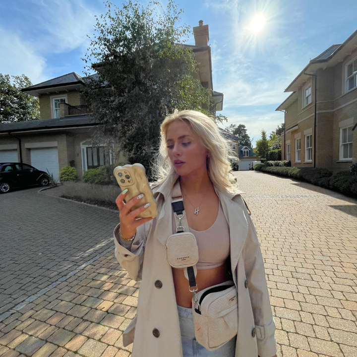 A woman with long blonde hair stands on a cobblestone driveway between two large houses under a bright, sunny sky. She is wearing a light-colored trench coat, a crop top, and ripped jeans. In one hand, she holds her iPhone15 adorned with Coconut Lane's 3D Pearl Phone Case - Gold and carries two small crossbody bags.