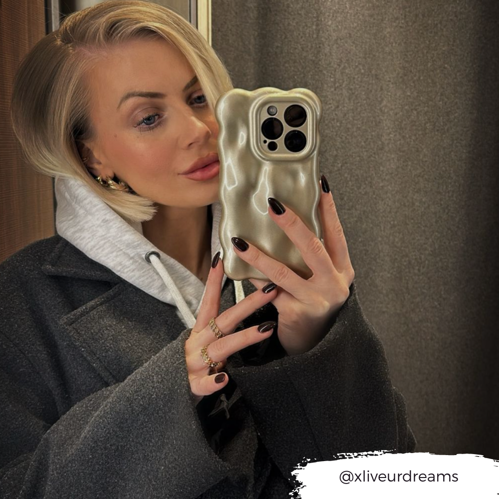 A person with short, blonde hair takes a mirror selfie with an iPhone15 encased in the Coconut Lane 3D Pearl Phone Case - Gold. They are wearing a dark overcoat layered over a light hoodie and showcasing well-manicured black nails. The Instagram handle @xliveurdreams is displayed in the bottom right corner.