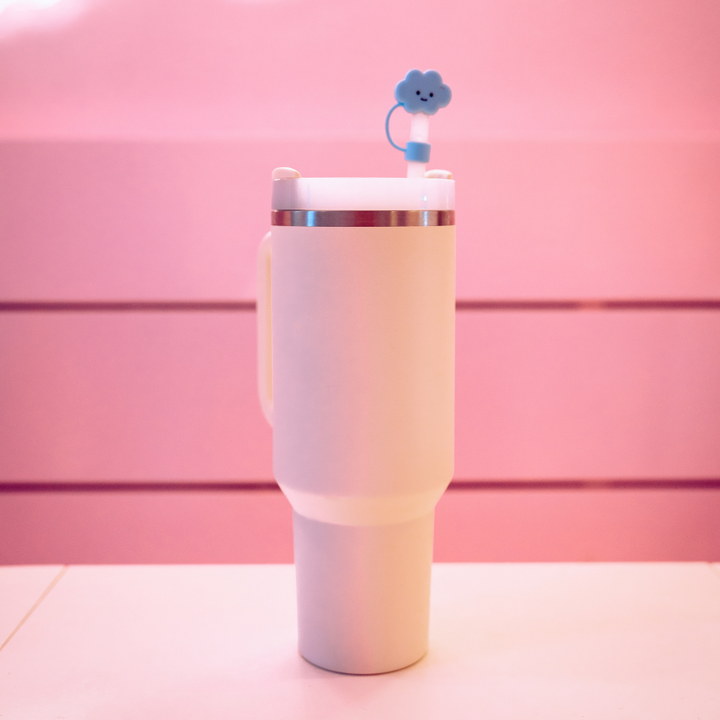 A white tumbler with a handle stands against a pink background. The lid is topped with Coconut Lane's Blue Cloud Straw Cover, featuring a happy face and crafted from high-quality silicone. This minimalist and playful design creates a cheerful atmosphere.