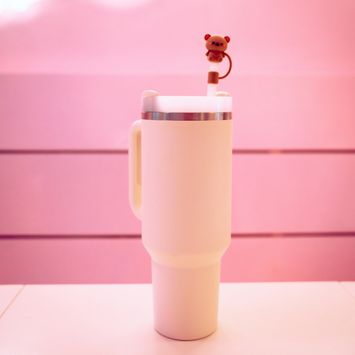 A white insulated travel mug with a lid and handle, featuring Coconut Lane's Teddy Bear Straw Cover on a straw, placed against a pastel pink background.