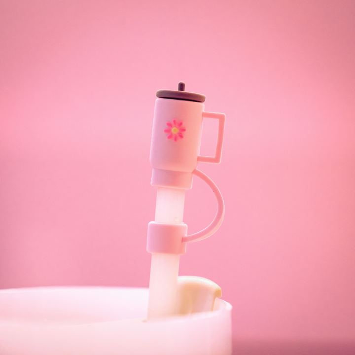 The Mini Pink Tumbler Straw Cover by Coconut Lane features a small, pink plastic coffee cup charm adorned with a tiny flower design, attached to a translucent silicone straw topper. The cohesive pastel aesthetic is complemented by a soft pink background.